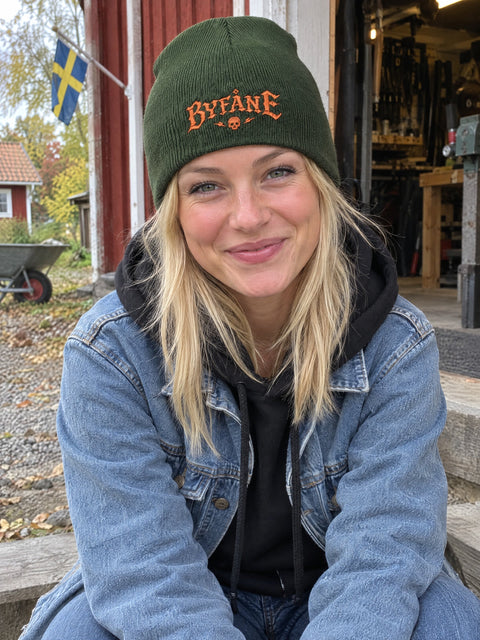 Byfåne på olivgrön Hatitude Beanie, buren av en yngre vuxen.
