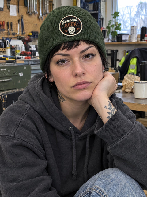 Some People Suck på olivgrön Hatitude Beanie, buren av en yngre vuxen.
